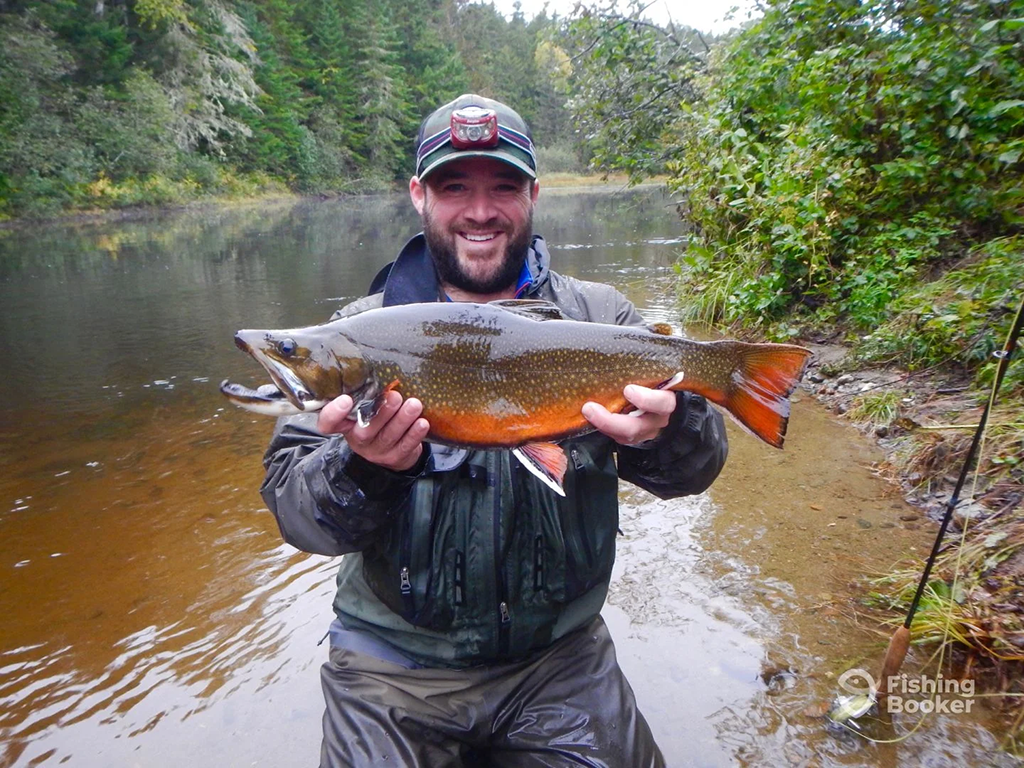 A man in waders stands by a riverbank, smiling at the camera as he holds a large Brook Trout. Behind him, you can see trees lining the water's edge and the gentle flow of the river.