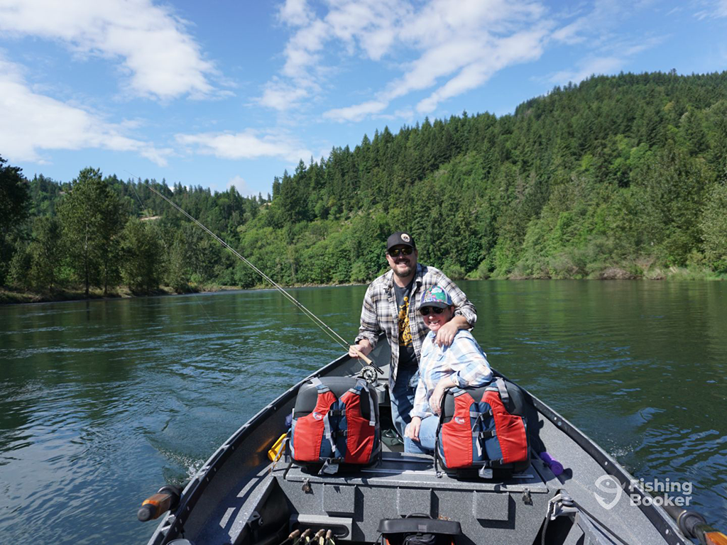 Two people sit in a small boat fishing on a calm river, surrounded by dense green forest under a blue sky. They've got fishing gear and backpacks with them in the boat.