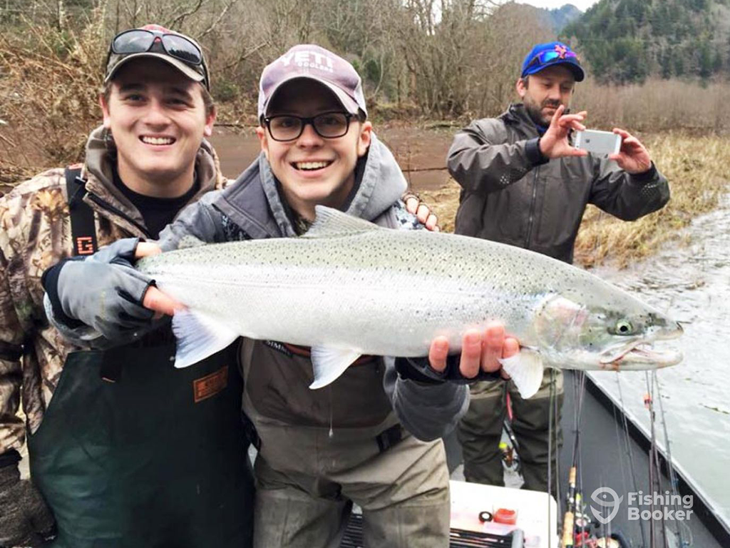 Two people on a boat in Oregon are smiling at the camera as they hold a large Steelhead, showing off their impressive catch. In the background, a third person snaps a picture with a smartphone to capture the memorable moment.