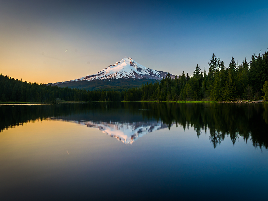 A snow-capped mountain reflected perfectly in a calm lake at sunset, surrounded by a dense forest—an idyllic spot in Oregon under a clear sky.