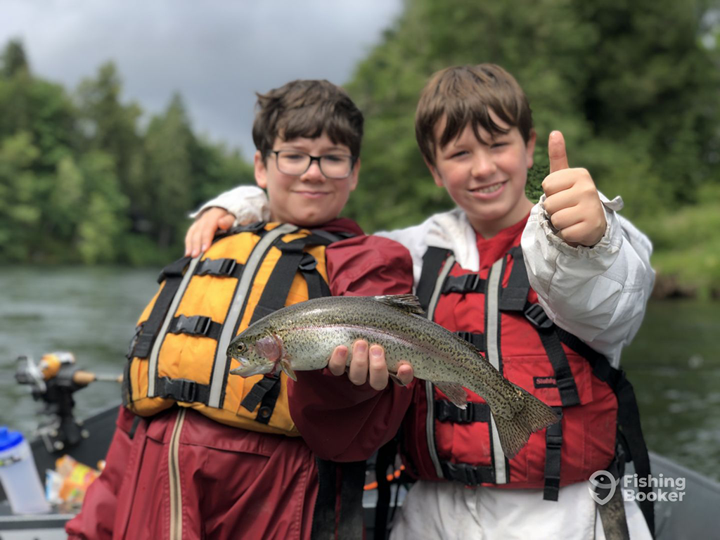 Two boys in life jackets on a boat, one proudly holding a Trout while the other gives a thumbs up.