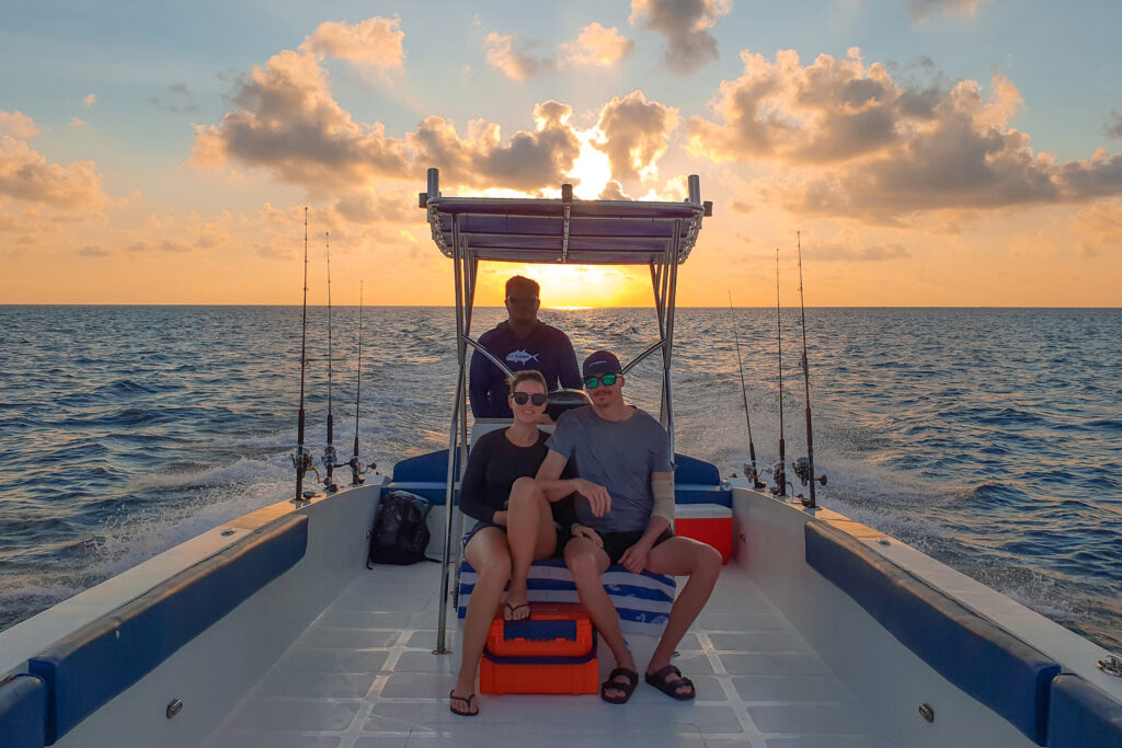Three people are riding on a small motorboat at sunset. Two sit at the front, wearing sunglasses, while the third stands behind them at the helm. Four fishing rods are mounted on the boat. In this tranquil scene, you might spot anglers hoping to catch popular species like Bass, Bluegill, or Trout. They could also be looking for larger catches such as Catfish or Northern Pike if they're cruising over deeper waters. For some, landing a Walleye or Perch would cap off a perfect evening on the lake. With four rods set up and dusk painting everything gold and orange, it's clear they’re ready for some serious fishing—and maybe even a memorable story about "the one that got away.