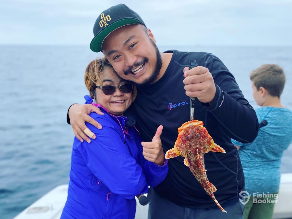 Two adults are smiling on a boat—one’s holding a Sculpin. The ocean stretches behind them, and another child is visible in the background.