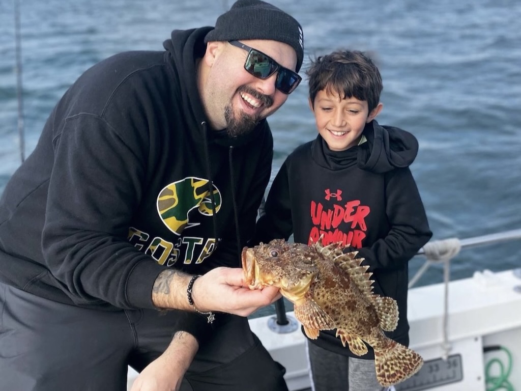 A man and a boy people on a boat, smiling at the camera as they hold a large, spiky fish. Water is visible in the background.