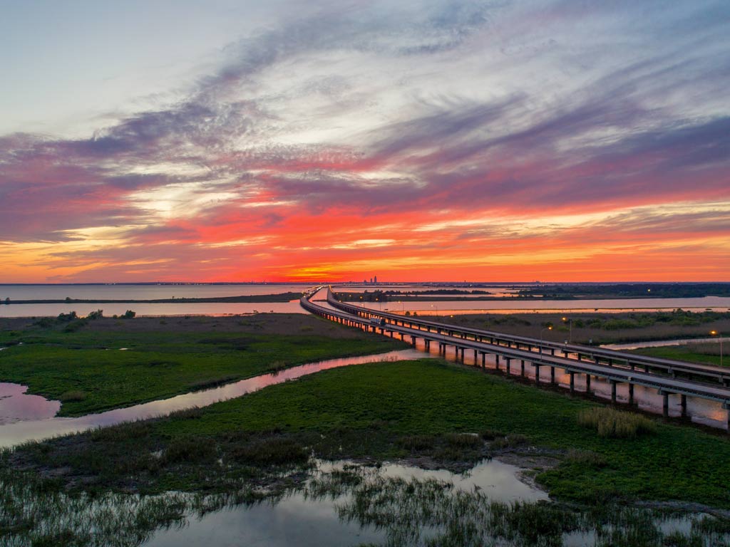 A sunset view of the Jubilee Parkway over Mobile Bay, Alabama, with swampy vegetation and waters surrounding the parkway .
