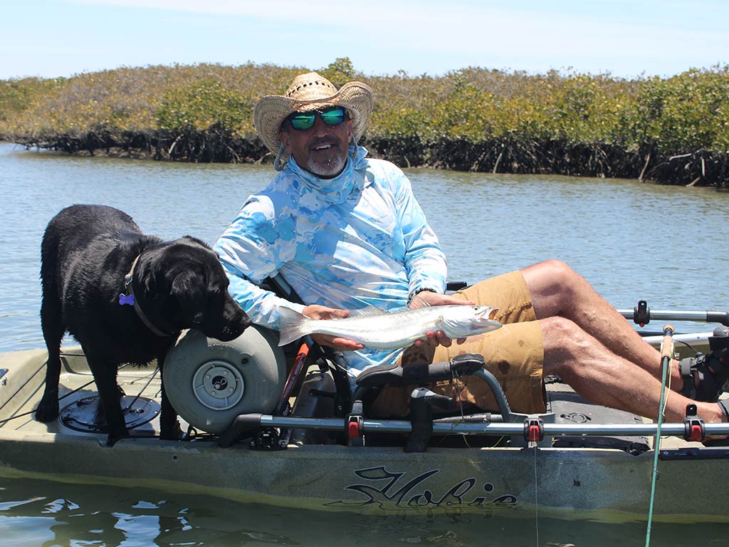 A man in a kayak holds a fish while a black dog beside him eagerly sniffs the catch. They’re floating on calm water near a wooded shoreline, surrounded by trees reflected on the lake’s surface, while fly fishing in Baja California.