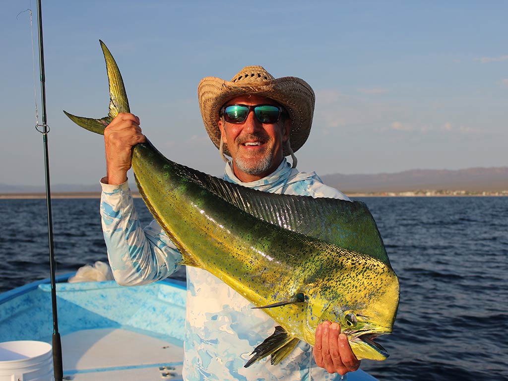 A man wearing a straw hat and sunglasses holds a large Mahi Mahi on a boat, with the ocean and distant shoreline in the background.