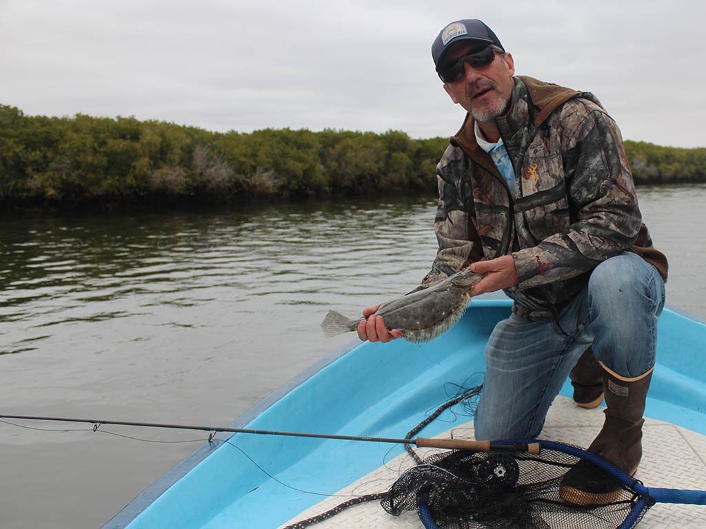 A man in outdoor gear kneels on a boat, holding a California Halibut beside a fishing net, showing off his Baja fly fishing skills with water and trees in the background.