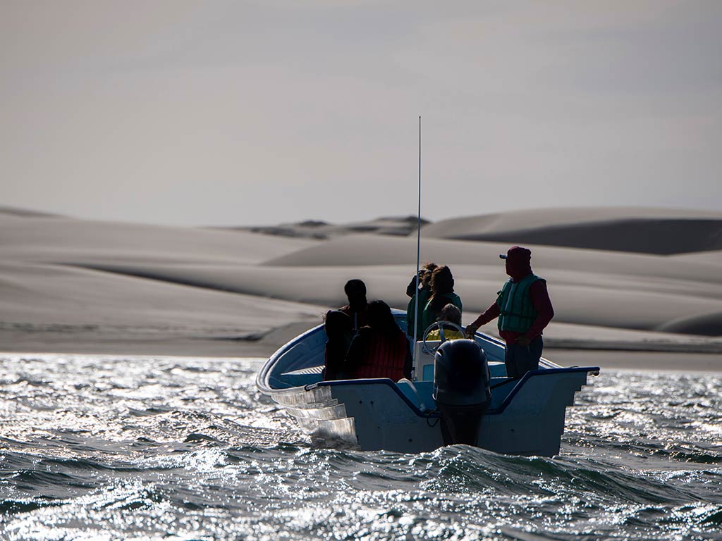 A view from behind of a small motorboat with four people in life jackets traveling across the water, with sandy dunes behind them, enjoying a bright sky, on Baja California's Magdalena Bay.