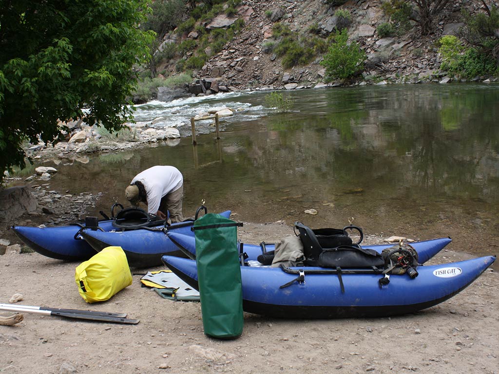 Two blue Inflatable Kayaks and gear sit on a riverbank, while a person bends over, getting equipment ready near the water. Trees and rocks fill the background.