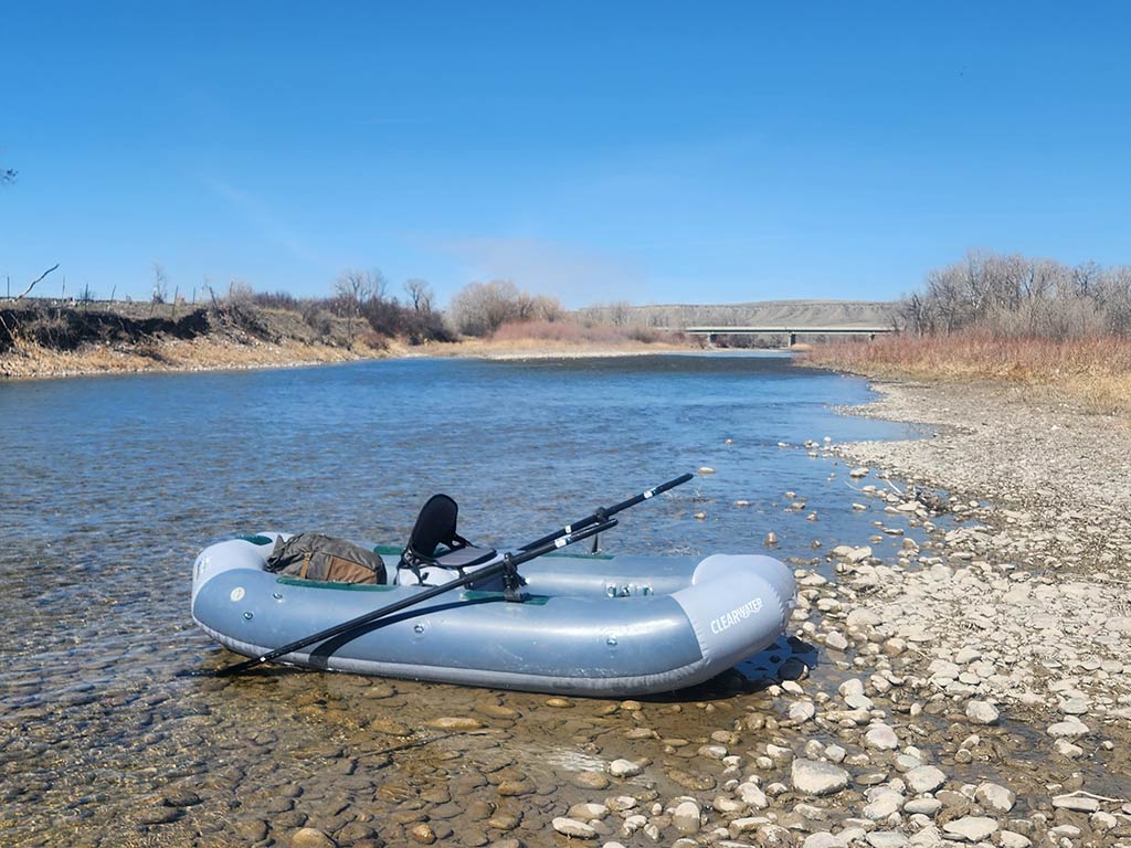 An inflatable float tube with oars rests on a rocky riverbank under a clear blue sky, with a bridge and trees in the background.