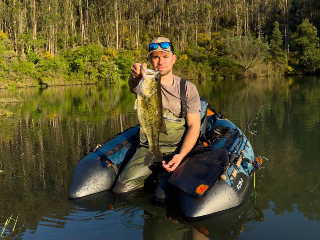 A man sitting in an inflatable float tube, fishing on a calm lake. He’s proudly holding up a large Bass in both hands. Behind him, lush trees and greenery line the shore, adding to the peaceful atmosphere.