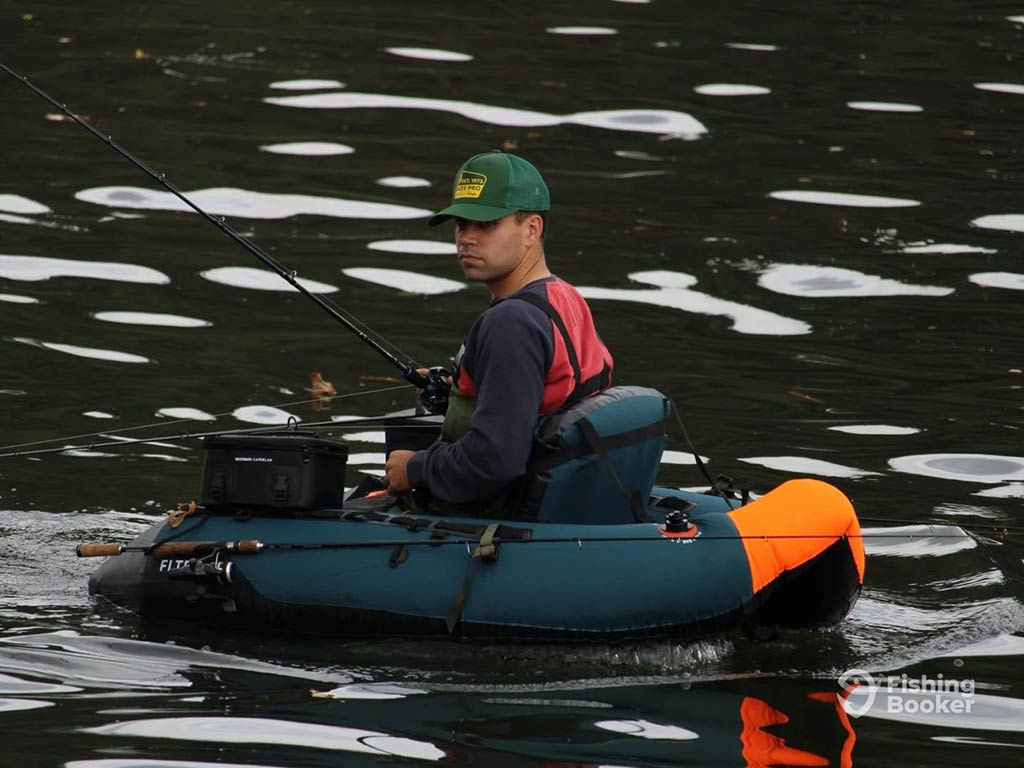 A man wearing a green cap sits in an inflatable float tube, enjoying fishing with his rod and tackle box on the water.