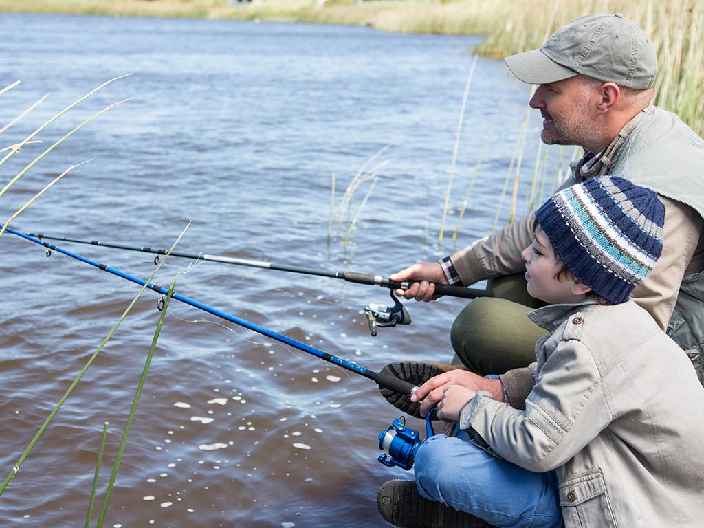 An adult and a child sit by the edge of a river, both holding fishing rods and looking at the murky water.