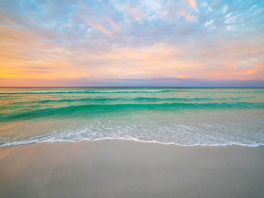 A calm ocean with gentle waves approaches a sandy beach under a colorful Fourth of July fishing sunset sky. Anglers might spot REDFISH, TARPON, and SNAPPER gliding nearshore as SEAGULLS soar overhead. The vibrant sky reflects on the water as everyone hopes to reel in a mighty FISH or two before dusk. It's an evening filled with anticipation—will you hook a SPECKLED TROUT, or maybe even a FLOUNDER? Either way, this peaceful moment on the sand promises memories as bright as the fireworks soon to light up the horizon.
