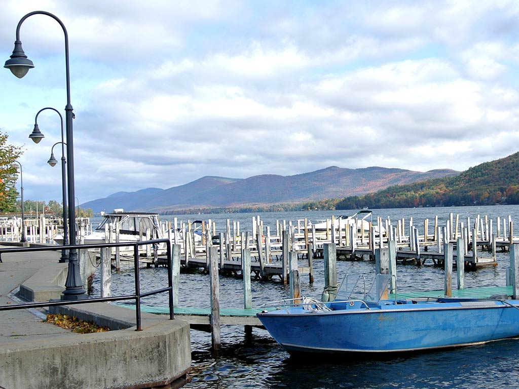 A blue boat docked near empty wooden piers on a lakeside, making it perfect for Fourth of July fishing. Lampposts line the walkway, and mountains rise under a cloudy sky. 