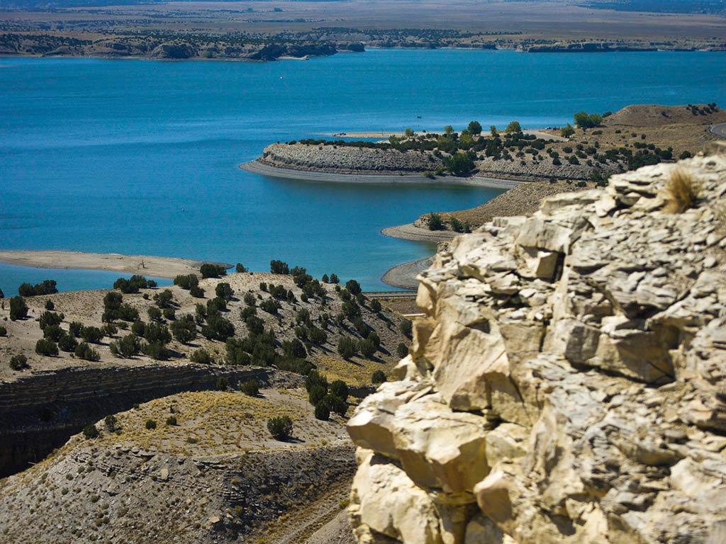 Rocky cliffs overlook a the blue waters of pueblo Reservoir, CO, with a curved shoreline. Scattered vegetation highlights the clear skies in this semi-arid landscape. 