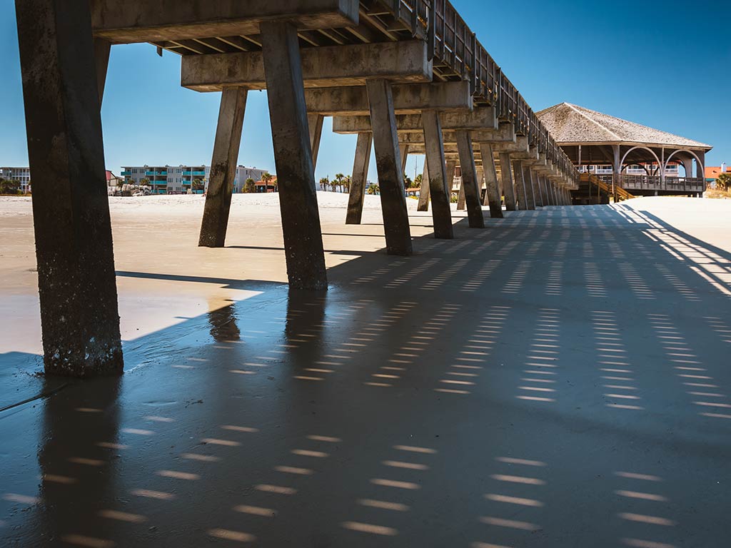 A pier stretches out over a sandy beach, casting rectangular shadows beneath it—perfect for Fourth of July fishing. There's a building at the end, and a clear blue sky overhead. Anglers might reel in Striped Bass, Flounder, or Bluefish from this spot, and you'll often see families enjoying the sunshine while waiting for their next catch. The gentle sound of waves mixes with the excitement each time someone lands a Mackerel or Porgy. It's the ideal place to spend an afternoon trying your luck, whether you're hoping for that big Black Sea Bass or just enjoying the view.