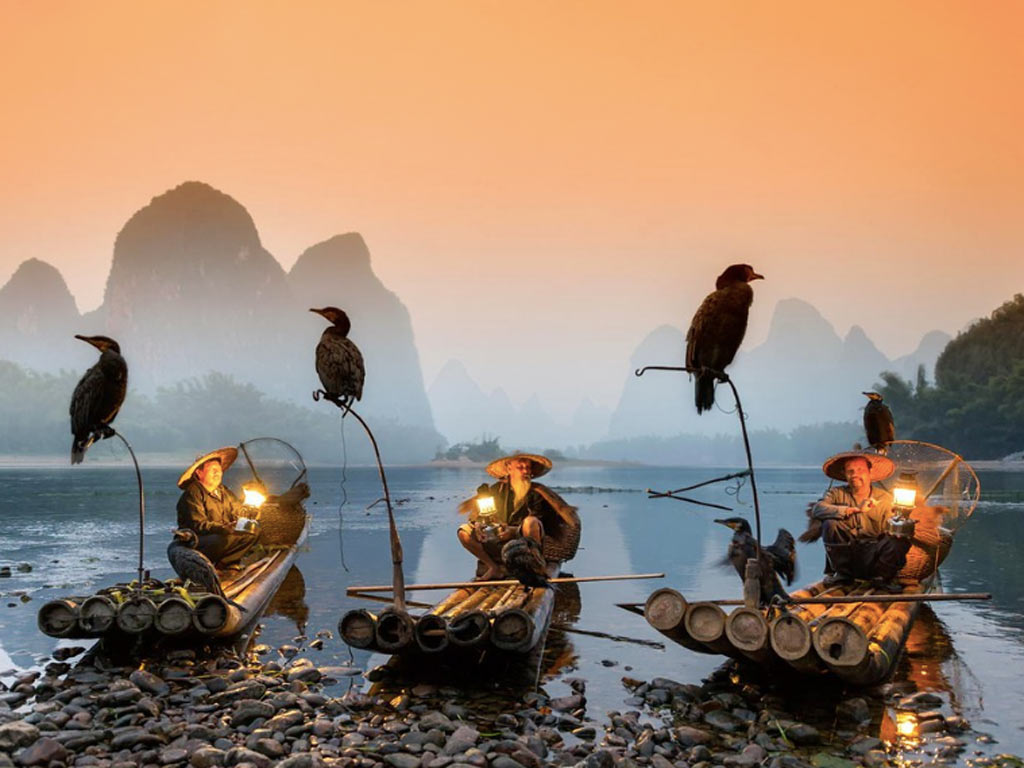 Three fishermen in traditional attire sit on bamboo rafts with cormorants and lanterns, set against a scenic river and mountain landscape at sunset.