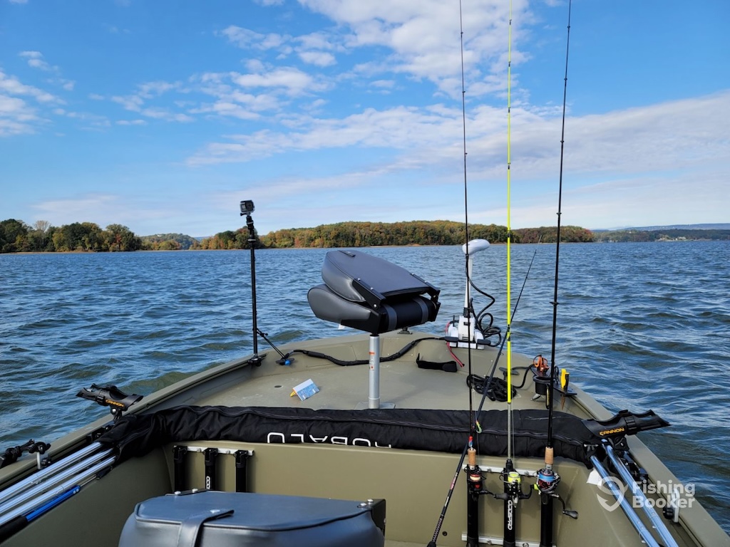 Two mounted seats face open water, while fishing rods and gear are neatly arranged and ready to go. A calm lake stretches out before the boat, with a distant tree-lined shore beneath partly cloudy skies.