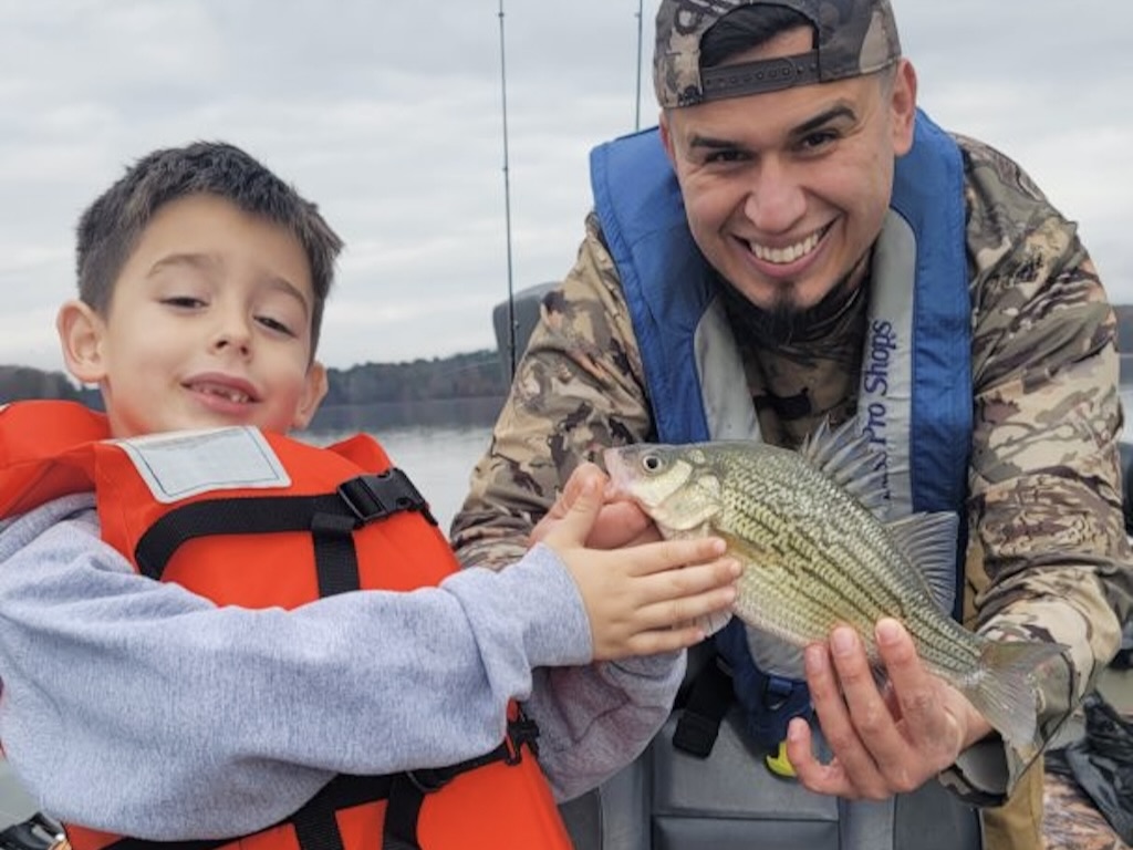 An adult and child, both wearing life jackets and camouflage, proudly hold a freshly caught Yellow Bass on a boat. Water and trees are visible in the background.