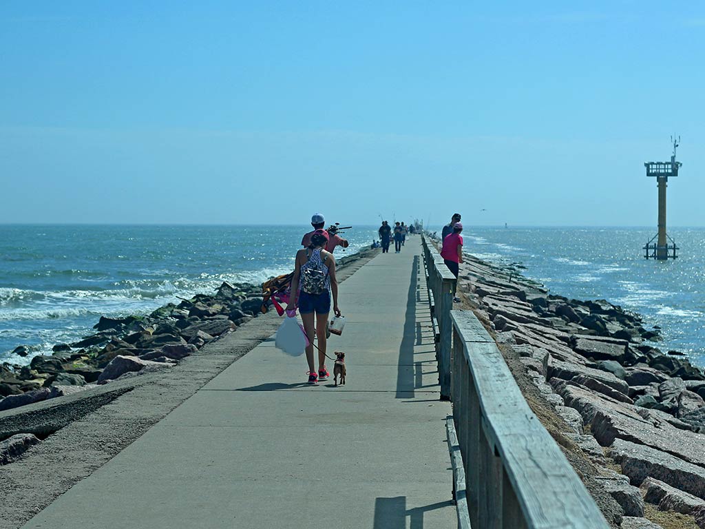 A view from behind as people walk along a concrete jetty in Freeport, Texas, surrounded by rocks and ocean, enjoying clear skies and perfect fishing tides near the distant structure.