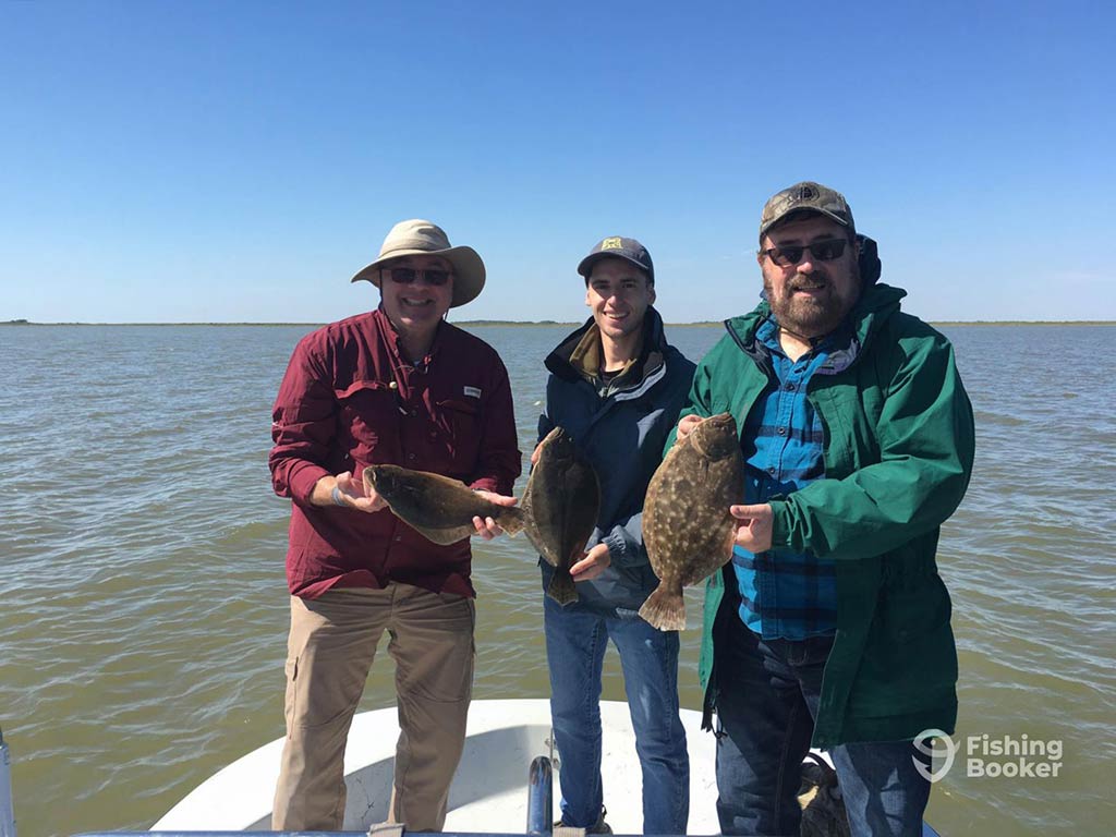 Three people on a boat in Freeport, Texas holding up Flounder they caught, with water and sky in the background.