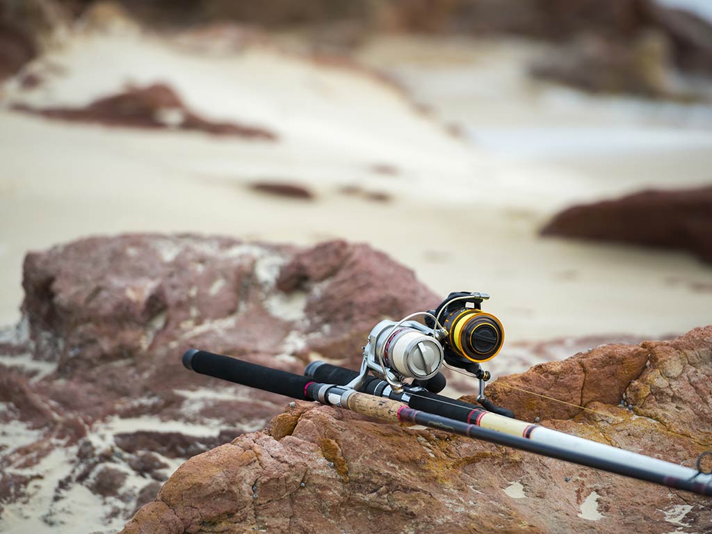 A fishing rod and reel rest on reddish rocks near a sandy shoreline.