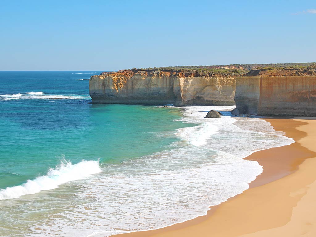 Coastal cliffs with a light brown rock face and turquoise water waves wash onto a sandy beach beneath a clear blue sky next to the Great Ocean Road in Victoria, Australia.