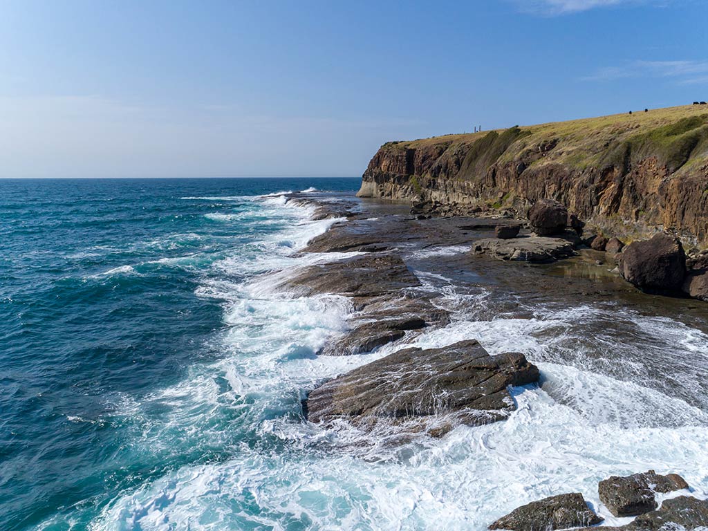 Waves crash against rocky cliffs under a clear blue sky, with grassy land above the shoreline and several people visible on the cliff edge near Illiwara.