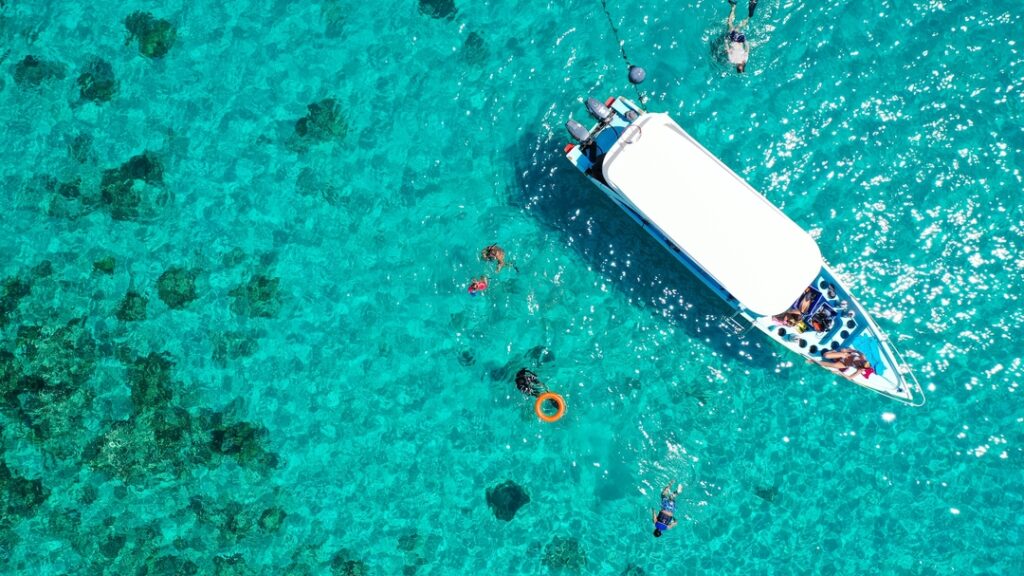 An aerial view shows a white boat floating on clear Turquoise water. Several people are swimming and snorkeling nearby, enjoying the vibrant marine life. The sunlight highlights the stunning shades of blue and green beneath them, making it easy to spot colorful species like Parrotfish, Angelfish, and Butterflyfish gliding around Coral Reefs below.