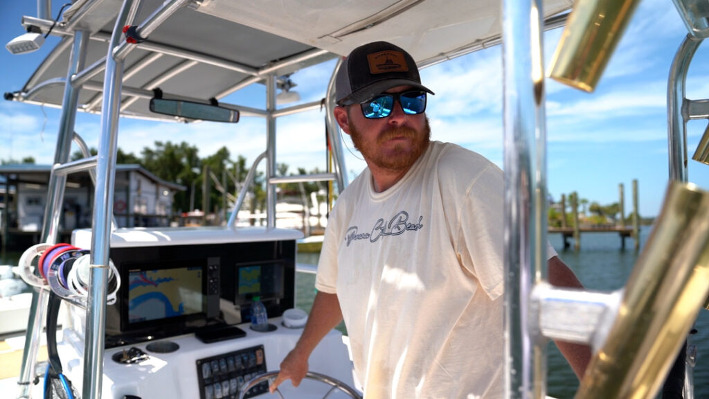 A man wearing sunglasses and a hat stands at the controls of a boat docked at a marina on a sunny day. The bright sunlight glints off the water as he scans the area, perhaps keeping an eye out for SNAPPER, GROUPER, or even MAHI-MAHI swimming near the docks. He's likely hoping he'll spot some REDFISH or SHEEPSHEAD close to the pilings, knowing they often hang around in search of crabs and shrimp. With his gear ready and rod nearby, he's eager to start his day's fishing adventure.