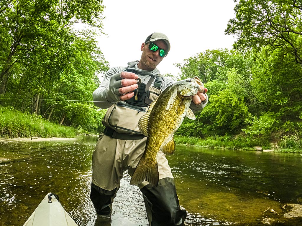 A man standing in a shallow creel, holding a large Bass proudly in both hands after fishing. He's dressed in waders and sunglasses, ready for fishing beneath the bright sky. Green trees surround the scene, their leaves reflecting off the water as he poses with his impressive catch.