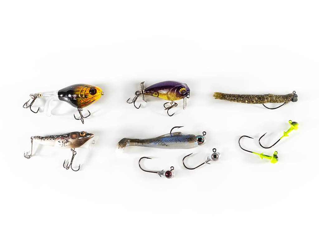 Six assorted fishing Lures and soft baits with hooks are arranged on a white background. The selection includes a minnow-shaped crankbait with vibrant green and yellow colors, a red crawfish plastic jig, a silver spoon lure with scale detailing, and a chartreuse worm soft bait.