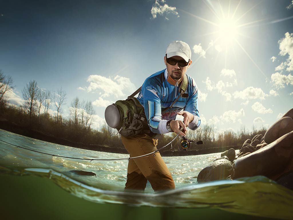A man wearing sunglasses and a cap is fly fishing in a creek under a sunny sky. There are trees and rocks in the background. 