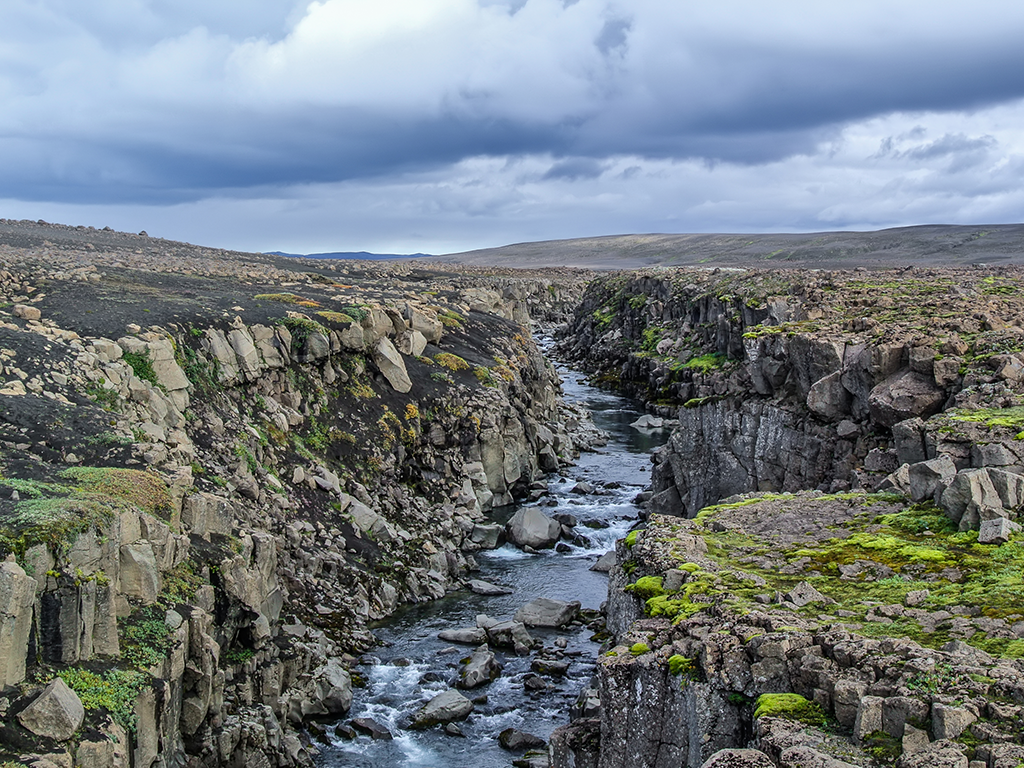 A rocky canyon with a river flowing through it with moss and sparse vegetation bordering the riverbanks, while a cloudy sky hangs overhead. 