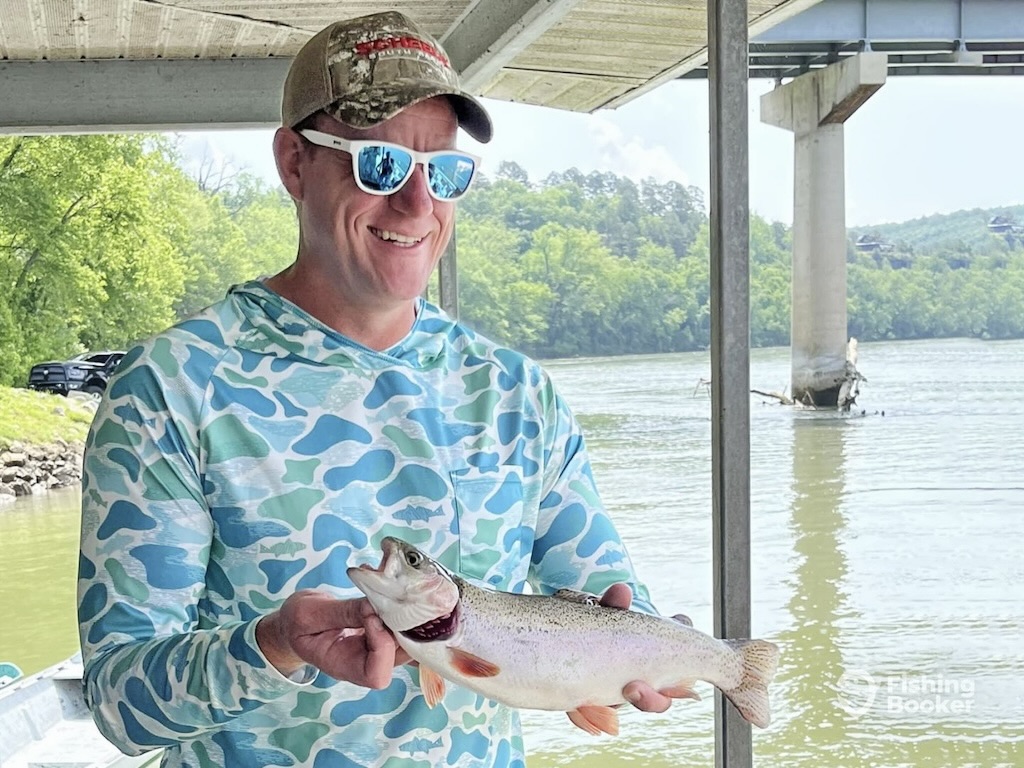 A man in a blue camouflage shirt and cap holds a Trout while fishing in Arkansas. He's standing on a dock by the river, with a bridge and trees visible in the background.
