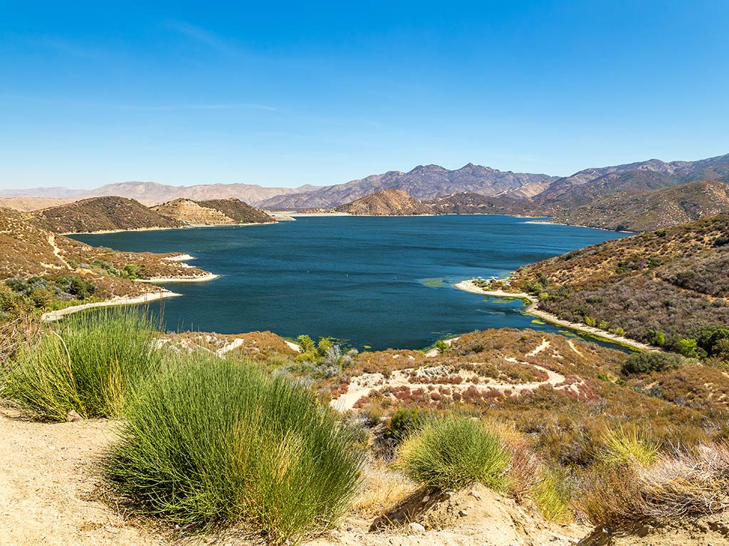 A clear blue lake is surrounded by dry, brown hills and sparse green vegetation under a bright, cloudless sky.