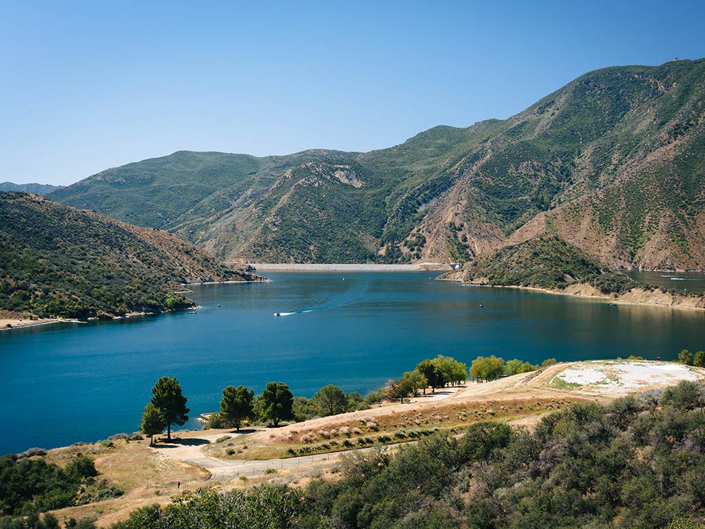 A large blue Lake Pyramid in California is surrounded by lush green hills and towering mountains. There’s a boat floating on the water under a clear, blue sky.