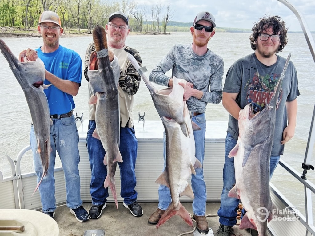 Four men on a boat proudly display Paddlefish they caught, with a lake and trees in the background.