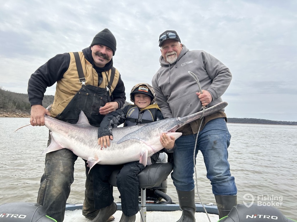 Three people on a boat are holding a large Paddlefish together, smiling at the camera under a cloudy sky, with calm water in the background.