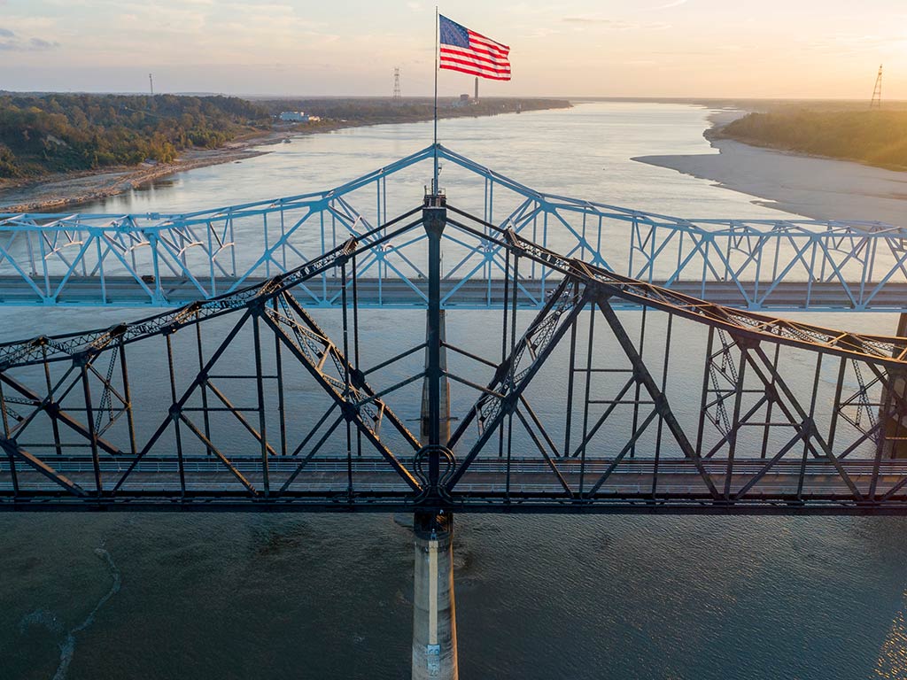 An American flag flies atop a metal bridge spanning the Mississippi River. Another bridge and forested banks appear in the background at sunset.