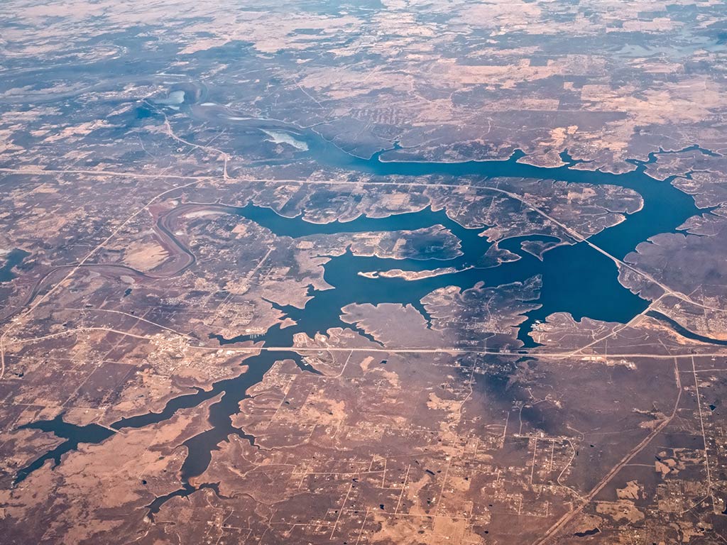 This aerial view shows a winding river cutting through a dry, sparsely vegetated landscape dotted with roads and scattered buildings. The landscape’s arid terrain provides little vegetation, highlighting the contrast between the blue-green riverbanks and the tan earth. 