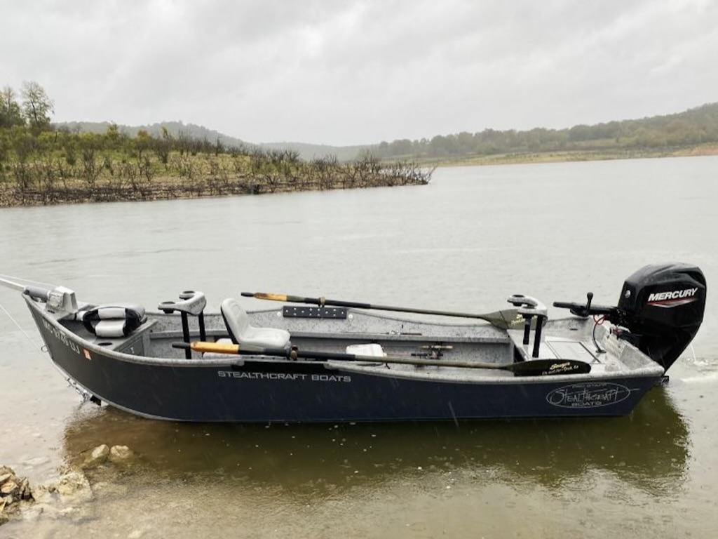 A black Stealthcraft fishing boat with a Mercury motor docked on the shore of Bull Shoals Dam on a cloudy day.