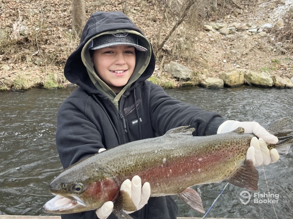 A child in winter fishing gear with their hood up holds up a large Rainbow Trout on the White River on a boat on a rainy day.