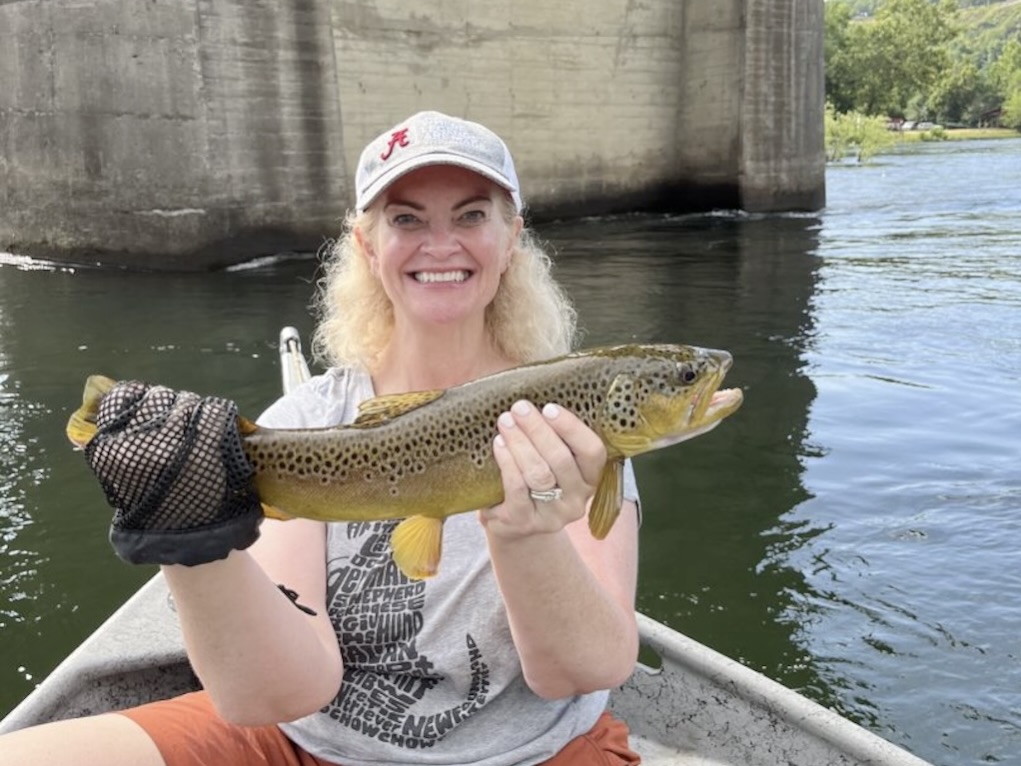 A woman with blonde hair in a baseball cap sitting on a boat next to Bull Shoals Dam in the White River on a bright day in Arkansas.
