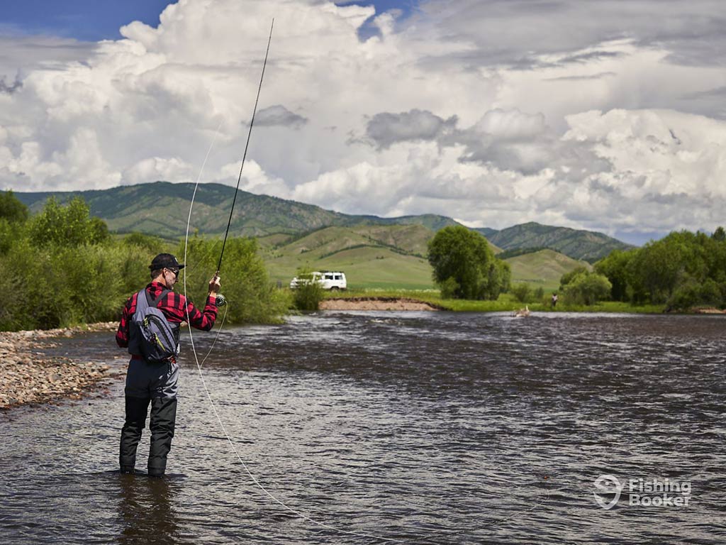 A person in fishing gear stands in a shallow river, casting a line for Taimen in Mongolia. Green hills and a white vehicle are visible in the background beneath a cloudy sky.