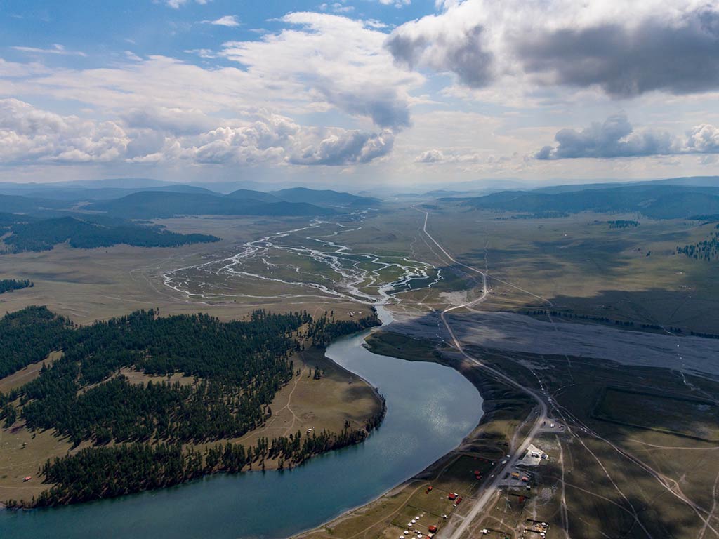 An aerial view shows the Eg River winding through a wide, green valley dotted with scattered trees and intersected here and there by roads&mdash;all beneath a partly cloudy sky in Mongolia.