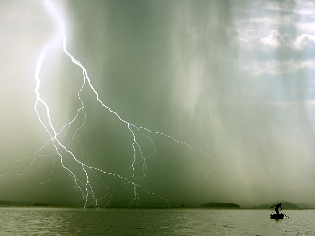 A view across a lake where a lone fishing boat is struggling to paddle away during a storm, with lightning visible on the left of the image.