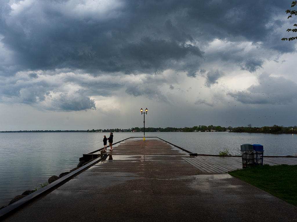 A view down a wet pier or jetty towards two people fishing under a cloudy, stormy sky.